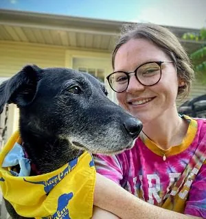 female with glasses wearing a tie dye shirt posing with a black dog wearing a yellow bandana around its neck